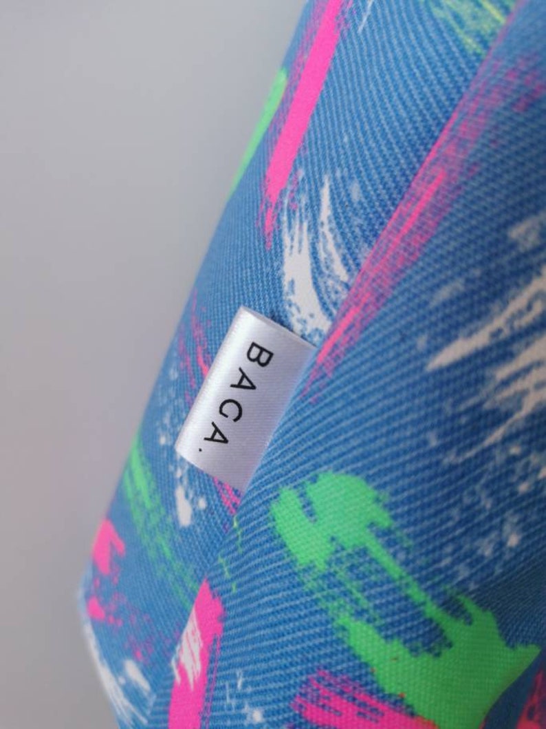 May include: A blue fabric surface featuring a textured pattern of diagonal brushstrokes in bright neon pink, lime green, and white. A small, rectangular white fabric tag is sewn onto the material, displaying the black text BACA. in a sans-serif font.