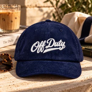 Puede incluir: Una gorra de béisbol de pana azul marino con el texto Off Duty bordado en blanco en una fuente cursiva en los paneles frontales. La gorra tiene una corona estructurada, una visera curva y costuras a juego en color azul marino.