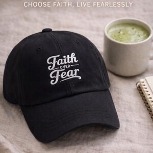 Puede incluir: Gorra de b&eacute;isbol negra con el texto bordado "Faith Over Fear" en escritura blanca. La imagen tambi&eacute;n incluye una taza de bebida verde y un peque&ntilde;o cuaderno, todo sobre una superficie de tonos neutros.