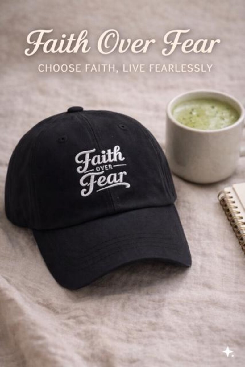 Puede incluir: Gorra de b&eacute;isbol negra con el texto bordado "Faith Over Fear" en escritura blanca. La imagen tambi&eacute;n incluye una taza de bebida verde y un peque&ntilde;o cuaderno, todo sobre una superficie de tonos neutros.