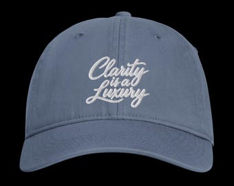 Peut inclure: Une casquette de baseball bleu ardoise avec une inscription brodée en blanc sur le panneau avant, lisant Clarity is a Luxury dans une police cursive. La casquette présente une calotte structurée, une visière courbée et des coutures.