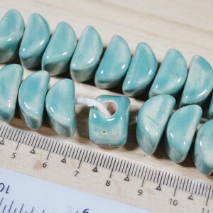 May include: A strand of teal ceramic beads, some shaped like half-moons and others square with a hole in the center. The beads are strung on a white thread and are lying on a wooden surface with a ruler.