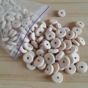 以下が含まれることがあります： さまざまなサイズのオフホワイトの木製ビーズの山。ビーズは丸く、中央に穴が開いています。ビーズはランダムなパターンで配置されています。