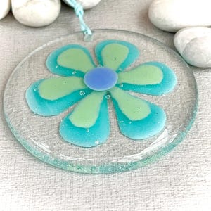May include: A circular glass ornament in the shape of a flower, with a light blue hanging string. The flower design features green and blue petals and a blue centre. The ornament is displayed on a white surface.