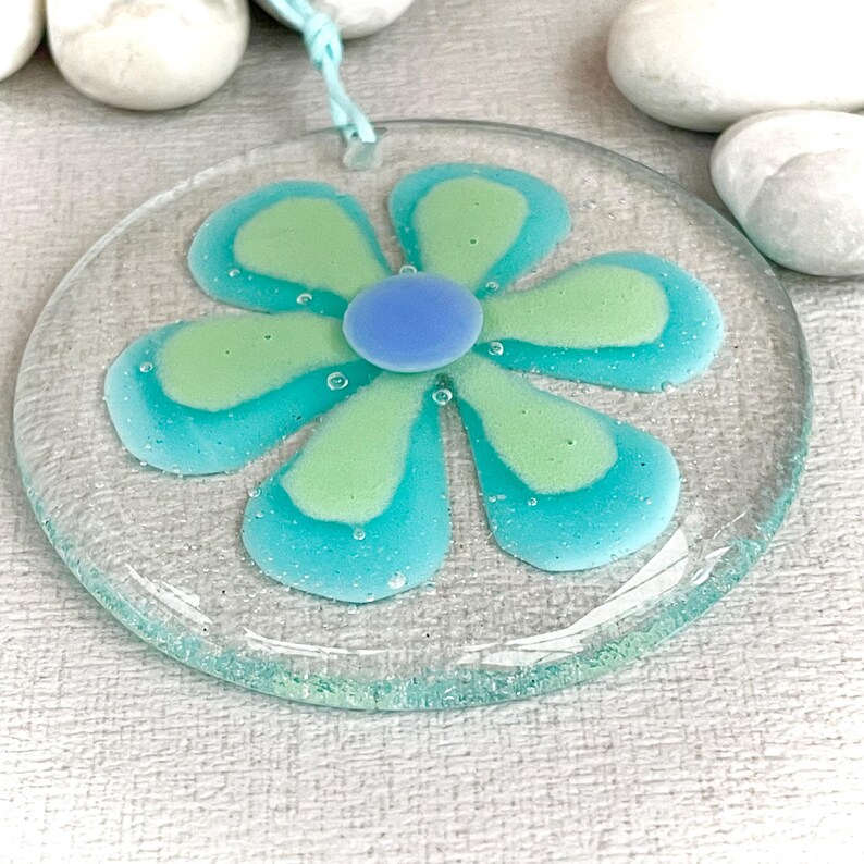 May include: A circular glass ornament in the shape of a flower, with a light blue hanging string. The flower design features green and blue petals and a blue centre. The ornament is displayed on a white surface.