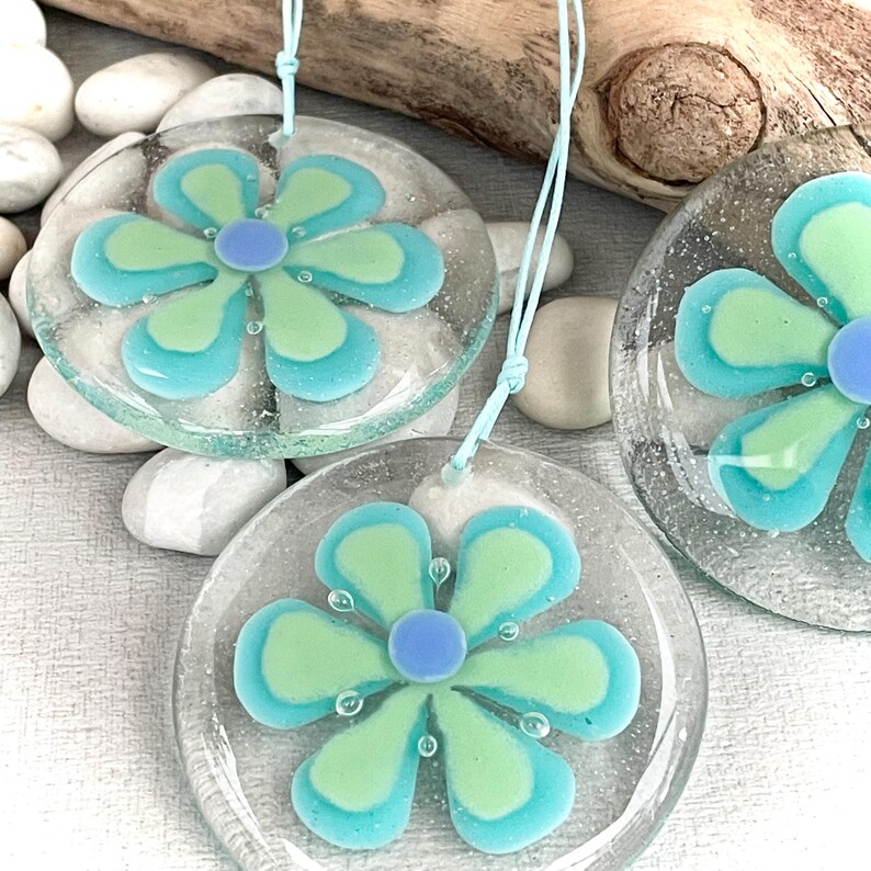 May include: Three clear glass flower ornaments with light blue string. Each ornament features a flower design with light green and turquoise petals and a blue centre. The ornaments are displayed on white stones.