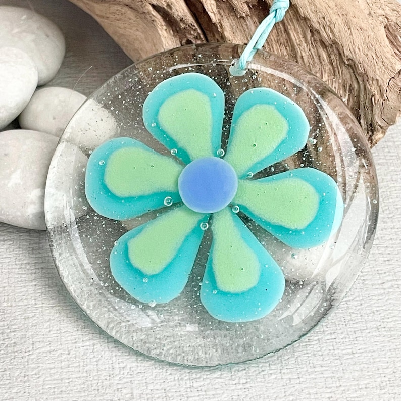 May include: A clear glass pendant featuring a flower design. The flower has light blue petals with a darker blue outline and a light green centre. A small blue bead is in the centre of the flower. A light blue cord is attached for hanging.