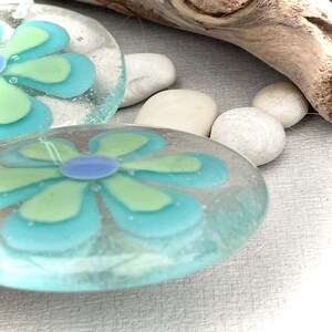 May include: Close-up of two clear glass flower-shaped art pieces with blue, light blue, and green petals. The centre of each flower is a small, round, light purple colour. White stones and a piece of driftwood are in the background.