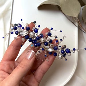 May include: A silver hair comb decorated with blue beads, clear crystals, and a large, faceted crystal. The comb has a delicate, floral design, suitable for weddings or special events. The comb is set against a white backdrop.