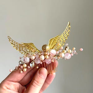 May include: A gold-colored hair comb featuring a decorative angel wing design. The comb is adorned with pink and clear crystals, pearls, and beads. The comb is held in a hand.