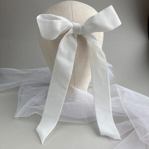 May include: A white satin ribbon bow with long tails, tied around a white mannequin head. The bow is displayed on a white tulle fabric.