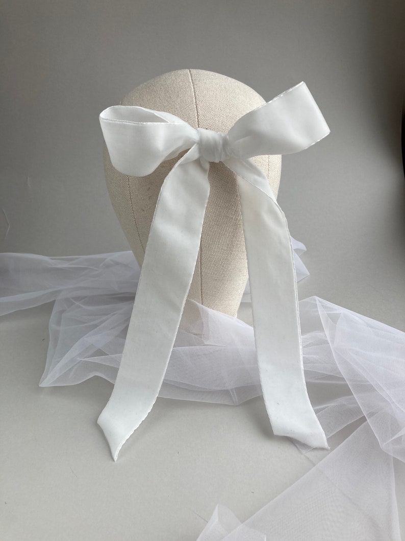 May include: A white satin ribbon bow with long tails, tied around a white mannequin head. The bow is displayed on a white tulle fabric.