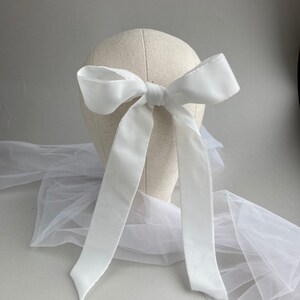 May include: A white satin ribbon tied into a large bow. The bow is attached to a white mannequin head. The ribbon is long enough to hang down the back of the head.