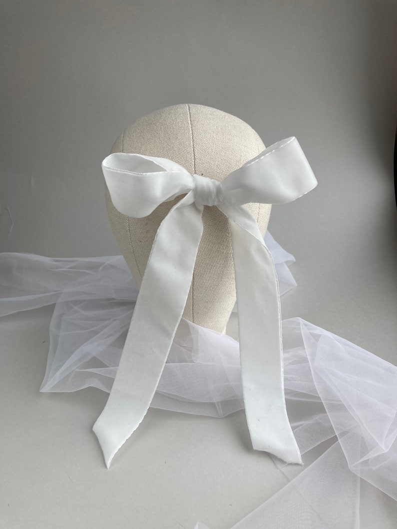 May include: A white satin ribbon tied into a large bow. The bow is attached to a white mannequin head. The ribbon is long enough to hang down the back of the head.