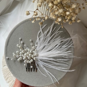 Könnte beinhalten: Ein weißer Federhaarkamm mit Perlenakzenten. Der Kamm liegt auf einer grauen Oberfläche.