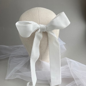 May include: A white satin ribbon tied into a large bow. The ribbon is long enough to be tied around a head or used as a hair accessory.