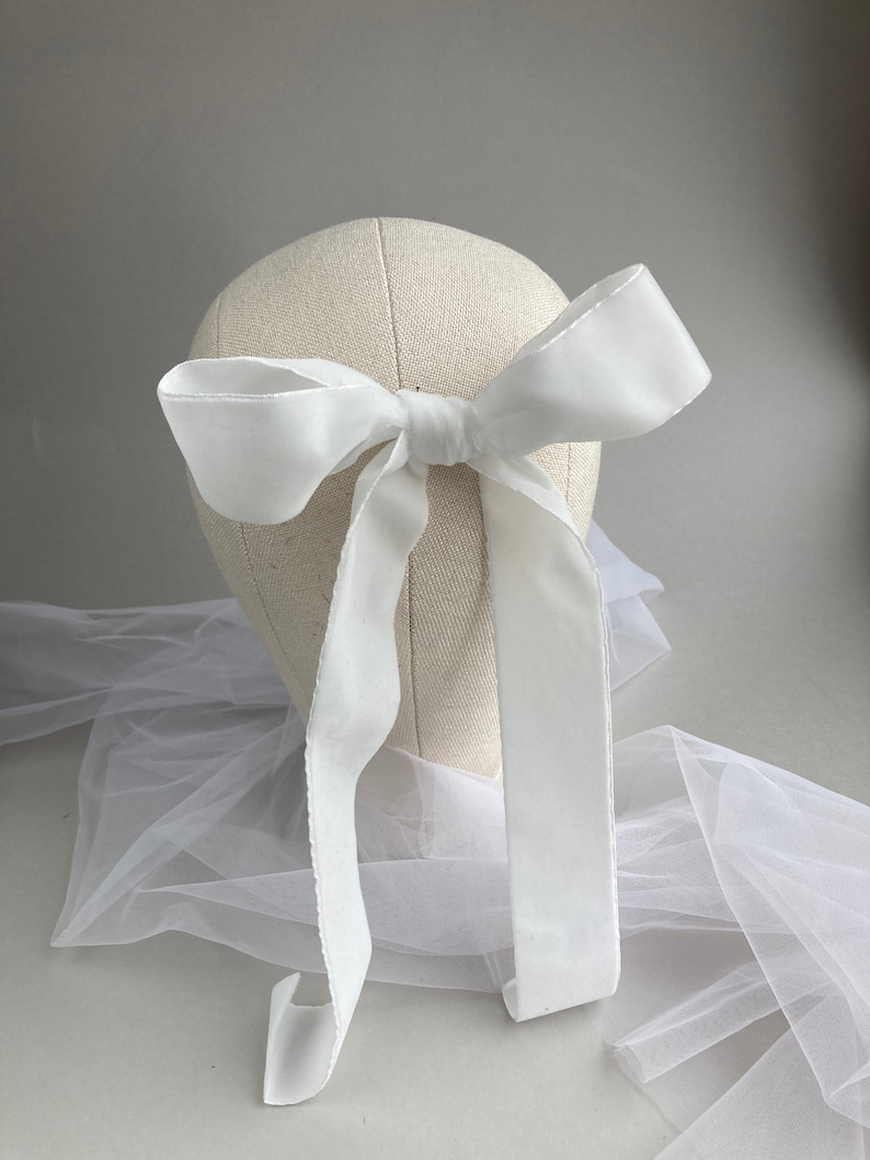 May include: A white satin ribbon tied into a large bow. The ribbon is long enough to be tied around a head or used as a hair accessory.
