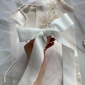 May include: A white satin ribbon tied into a bow. The ribbon is held in a hand against a white background.