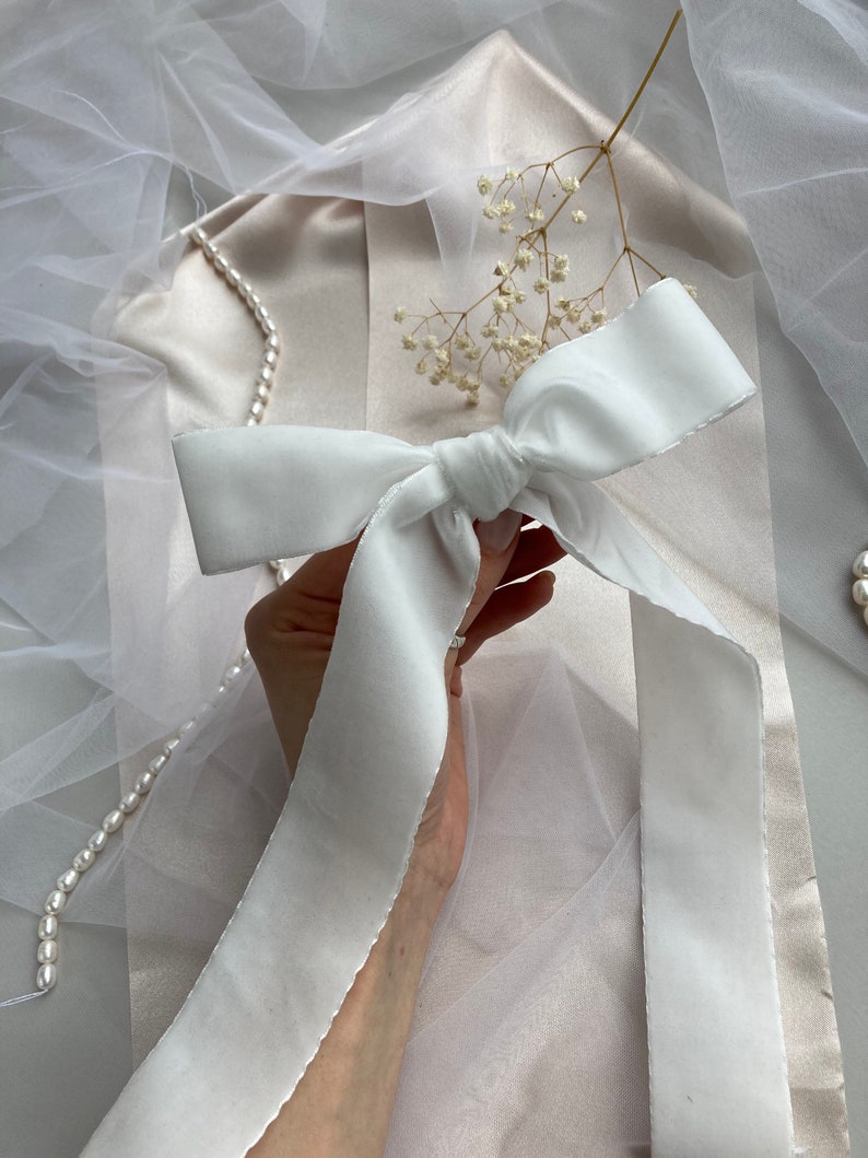 May include: A white satin ribbon tied into a bow. The ribbon is held in a hand against a white background.