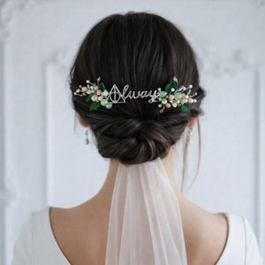 May include: A decorative hair comb with the word "Always" and a triangle symbol, adorned with pink and green beads and leaves. The comb is set against dark hair, with a white veil and dress visible.
