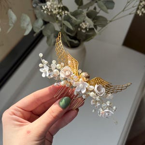 May include: A gold-colored hair comb with a decorative angel wing design. The comb features white flowers, pearls, and clear crystals. The wings are made of a delicate, openwork metal. A beautiful hair accessory for special occasions.