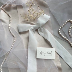 May include: White satin ribbon tied into a bow with long tails. The ribbon is on a light brown surface with a white tulle fabric and a small bouquet of white flowers. A white card with the text "Spring Wind accessories" is also on the surface.