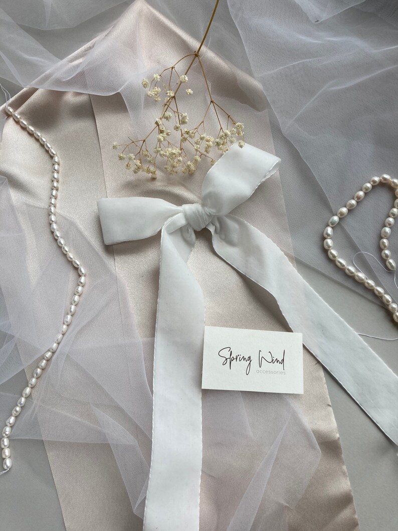 May include: White satin ribbon tied into a bow with long tails. The ribbon is on a light brown surface with a white tulle fabric and a small bouquet of white flowers. A white card with the text "Spring Wind accessories" is also on the surface.