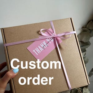 May include: A brown cardboard box tied with a light pink ribbon and a matching tag that reads "Thank you for your order". The words "Custom order" are printed in white on the box. The box is on a white background.