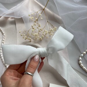 May include: White satin ribbon tied into a bow with a silver trim. The ribbon is on a light pink surface with white flowers and a white sheer fabric.