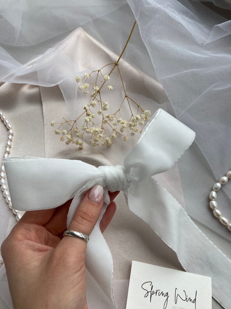 May include: White satin ribbon tied into a bow with a silver trim. The ribbon is on a light pink surface with white flowers and a white sheer fabric.