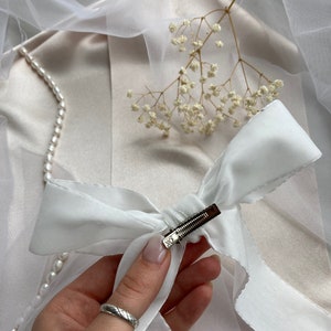 May include: A white satin ribbon bow with a silver clip. The bow is tied in a simple knot and the clip is attached to the center of the bow.