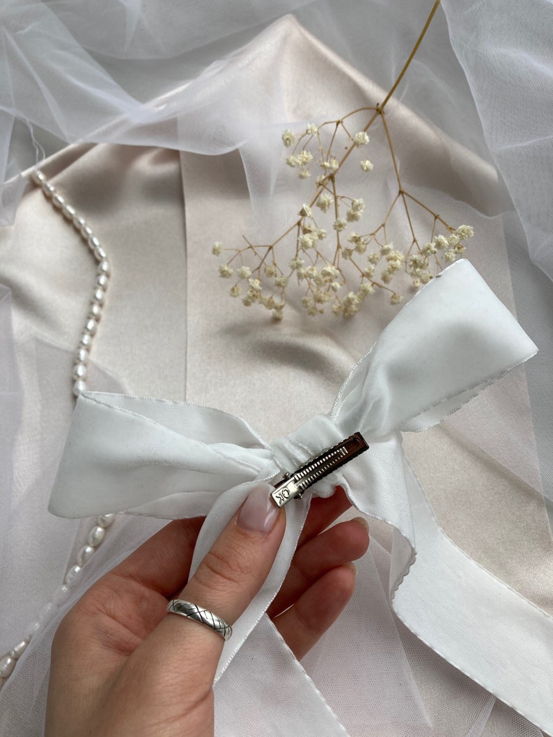 May include: A white satin ribbon bow with a silver clip. The bow is tied in a simple knot and the clip is attached to the center of the bow.