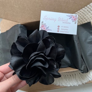 Może przedstawiać: Duży czarny kwiat z satyny z wieloma warstwami płatk&oacute;w. Kwiat znajduje się w pudełku z wizyt&oacute;wką, na kt&oacute;rej widnieje napis "Spring Wind accessories" oraz numer telefonu, ikona Facebooka i ikona Instagrama.