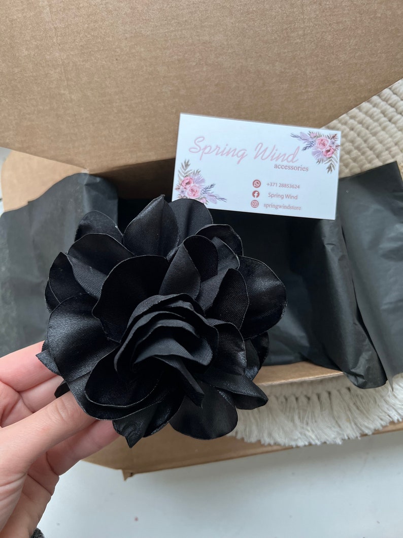 Może przedstawiać: Duży czarny kwiat z satyny z wieloma warstwami płatk&oacute;w. Kwiat znajduje się w pudełku z wizyt&oacute;wką, na kt&oacute;rej widnieje napis "Spring Wind accessories" oraz numer telefonu, ikona Facebooka i ikona Instagrama.
