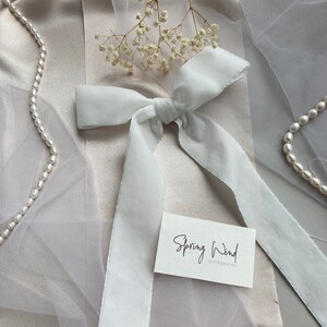 May include: White satin ribbon tied into a bow on a light beige fabric background. The ribbon is long and flowing. A small white card with the text "Spring Wind accessories" is on the fabric.