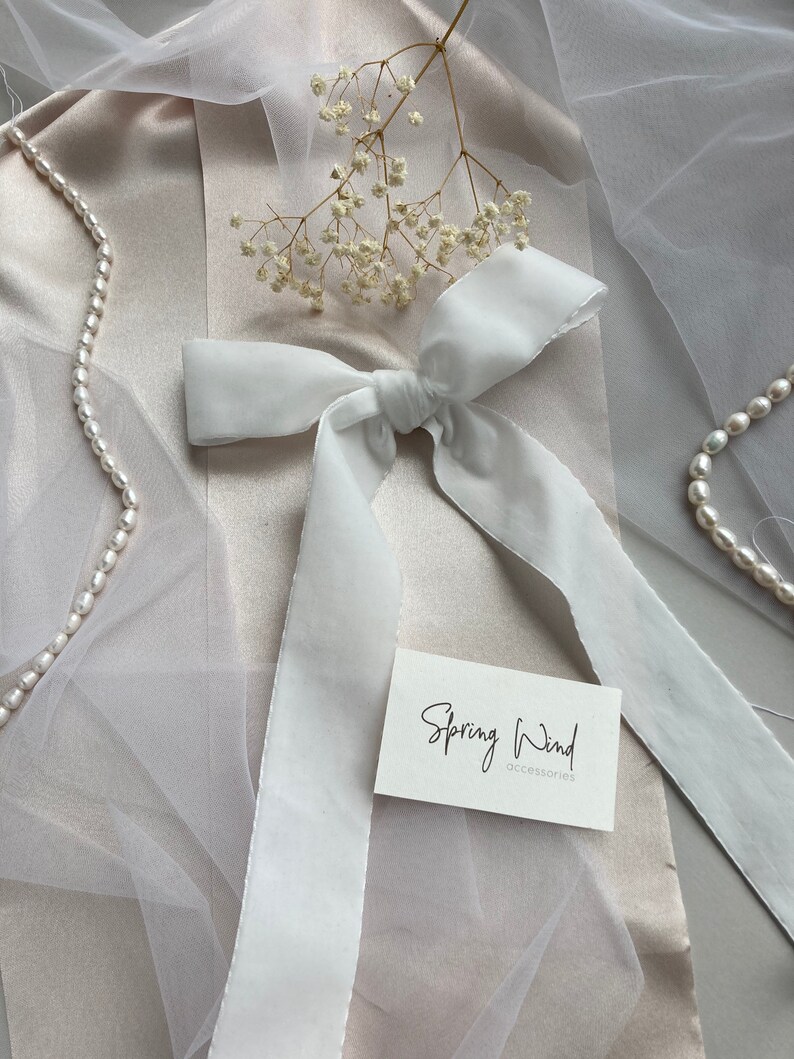 May include: White satin ribbon tied into a bow on a light beige fabric background. The ribbon is long and flowing. A small white card with the text "Spring Wind accessories" is on the fabric.