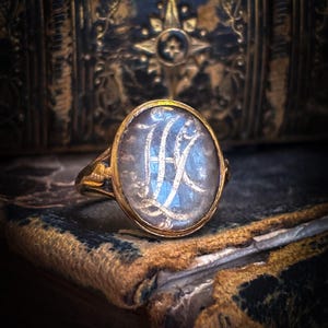 May include: A gold ring with an oval, iridescent stone featuring a white monogram. The ring is set against a backdrop of a vintage, leather-bound book with gold accents. The ring's band has ornate detailing.