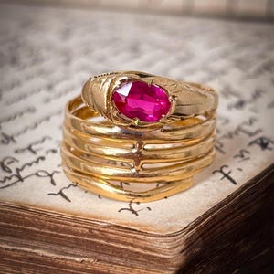 May include: A gold ring featuring a vibrant oval-cut pinkish-red gemstone set in a textured band. The ring consists of multiple stacked, interconnected gold bands that create a layered appearance, with the primary stone centered at the top.