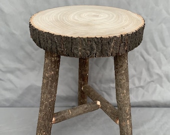 May include: A rustic wooden stool with a round, natural wood top and three sturdy legs. The top has a bark-like texture and the legs are made of rough-hewn branches.