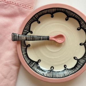 Può includere: Un piccolo piatto decorativo in ceramica con un cucchiaio abbinato. Il piatto ha un bordo rosa e un centro color crema, con un bordo a motivi bianchi e neri. Il cucchiaio è rosa con un manico a righe bianche e nere. Misure in centimetri.