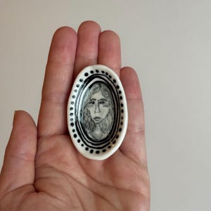 May include: Oval-shaped ceramic dish with a hand-drawn portrait of a person. The portrait is in black and white, framed by black lines and dotted border. The dish is held in a hand.