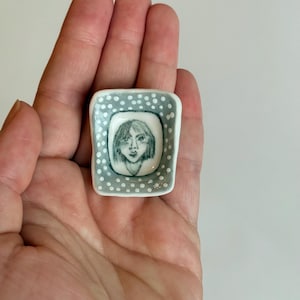 May include: A small, square ceramic dish with a hand-drawn portrait of a person. The portrait is in black and white, set against a white background. The dish has a gray dotted border. The dish is held in a hand.