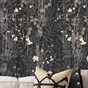 Op de afbeelding: Donker behang met een verweerd houteffect, met een botanisch patroon van witte bessen, grijze bladeren en beige bloemen. Het ontwerp bevat varens en ander gebladerte. De onderkant van de afbeelding toont witte bankkussens en decoratieve kussens.