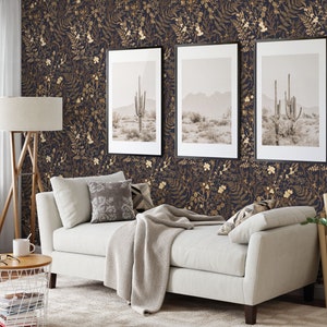 May include: A living room interior with a white sofa, a wooden floor, and a dark blue wallpaper with gold floral patterns. Three framed pictures of a desert landscape with cacti hang on the wall. A white side table with a wire basket and books sits next to the sofa.