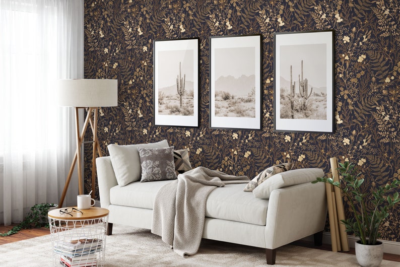May include: A living room interior with a white sofa, a wooden floor, and a dark blue wallpaper with gold floral patterns. Three framed pictures of a desert landscape with cacti hang on the wall. A white side table with a wire basket and books sits next to the sofa.