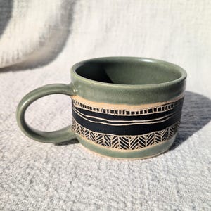 May include: A handmade ceramic mug with a green glaze. The mug features a black and beige band with a geometric design. The handle is also green. The mug is sitting on a textured, light gray surface.