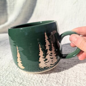 May include: A dark green ceramic mug with a rounded shape and a large handle. The mug features a design of three stylized trees in a lighter, tan color. The mug is designed for hot beverages.