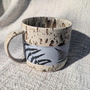 May include: A ceramic mug with a speckled beige and brown exterior. A light blue band encircles the mug, featuring black fern leaf illustrations. The mug has a curved handle and is suitable for hot or cold beverages.