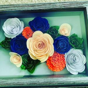 May include: A framed paper flower arrangement featuring a variety of colours, including white, blue, orange, and peach. The flowers are arranged in a cluster and are surrounded by green leaves.