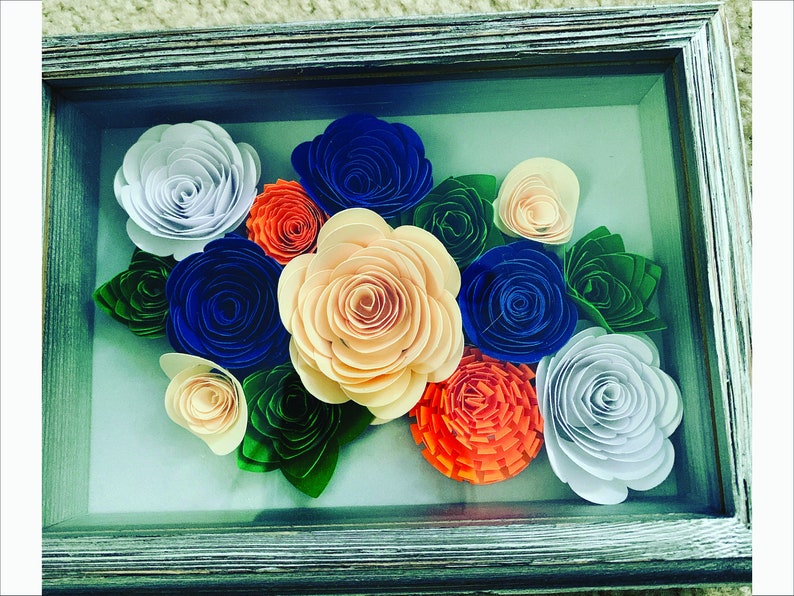 May include: A framed paper flower arrangement featuring a variety of colours, including white, blue, orange, and peach. The flowers are arranged in a cluster and are surrounded by green leaves.